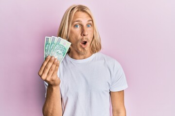 Caucasian young man with long hair holding 100 polish zloty banknotes scared and amazed with open mouth for surprise, disbelief face