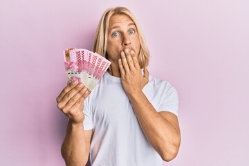 Caucasian young man with long hair holding 100000 indonesian rupiah covering mouth with hand,...