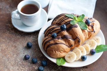 Croissant with chocolate, blueberries, bananas, and mint on a dark brown background. Breakfast with a croissant and a cup of coffee