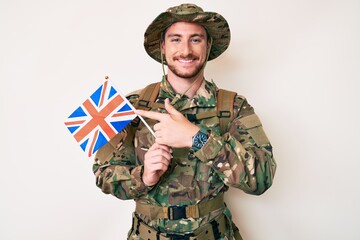 Young caucasian man wearing camouflage army uniform holding united kingdom flag smiling happy pointing with hand and finger