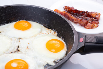 A nutritious breakfast of fried eggs and crispy bacon. The process of cooking a group of eggs in a pan. Crispy bacon on a plate. Idea for a quick, hearty American or British breakfast.