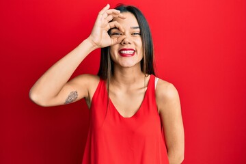 Young hispanic girl wearing casual style with sleeveless shirt doing ok gesture with hand smiling, eye looking through fingers with happy face.