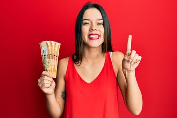 Young hispanic girl holding 500 philippine peso banknotes smiling with an idea or question pointing finger with happy face, number one