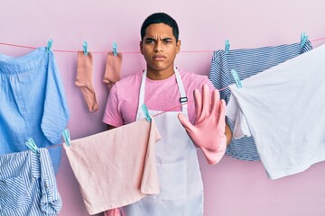 Young handsome hispanic man wearing cleaner apron holding clothes on clothesline doing stop sing...