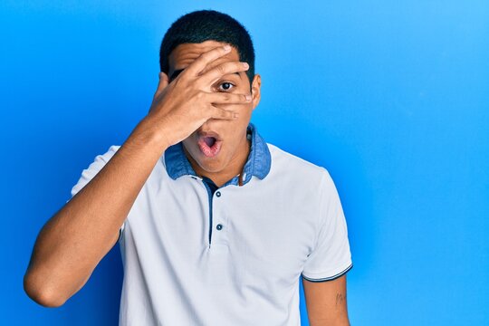 Young handsome hispanic man wearing casual clothes peeking in shock covering face and eyes with hand, looking through fingers afraid