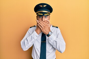 Handsome hispanic man wearing airplane pilot uniform shocked covering mouth with hands for mistake. secret concept.