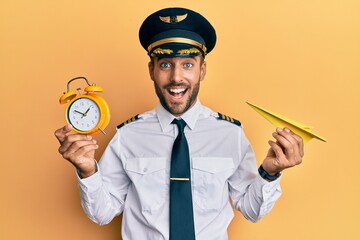 Handsome hispanic pilot man holding paper plane and alarm clock smiling and laughing hard out loud...