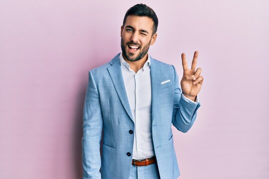 Young hispanic businessman wearing business jacket smiling with happy face winking at the camera doing victory sign. number two.
