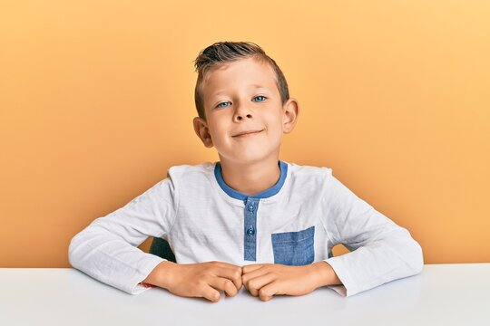 Adorable caucasian kid wearing casual clothes sitting on the table relaxed with serious expression on face. simple and natural looking at the camera.