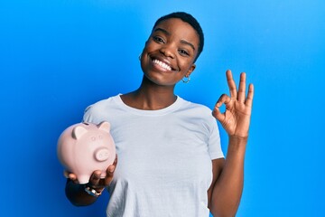 Young african american woman holding piggy bank doing ok sign with fingers, smiling friendly...