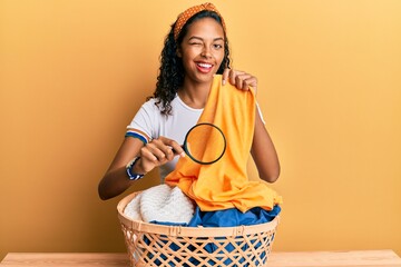 Young african american girl holding magnifying glass looking for stain at clothes winking looking...