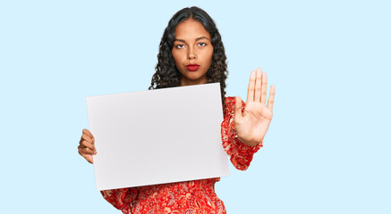 Young african american girl holding blank empty banner with open hand doing stop sign with serious...