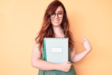 Young beautiful woman wearing glasses holding book pointing finger to one self smiling happy and proud