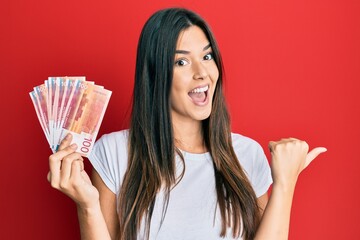 Young brunette woman holding 100 norwegian krone banknotes pointing thumb up to the side smiling...