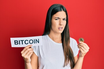Young brunette woman holding bitcoin virtual money clueless and confused expression. doubt concept.
