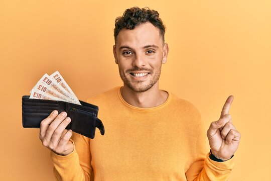 Young hispanic man holding wallet with united kingdom pounds smiling happy pointing with hand and finger to the side