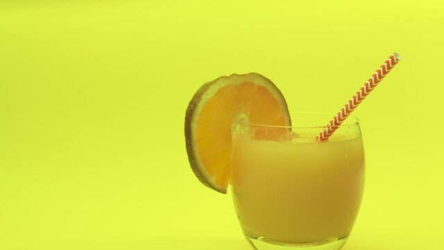 Pour yellow lemonade on a bright yellow background. Orange juice in a glass, studio shot. Fruit alcohol. Cocktail glass with straw and lime wedge