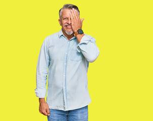 Middle age grey-haired man wearing casual clothes covering one eye with hand, confident smile on face and surprise emotion.