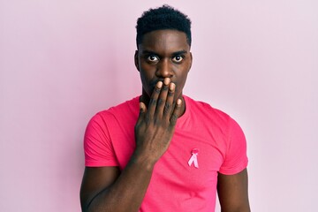 Handsome black man wearing pink cancer ribbon attached on shirt covering mouth with hand, shocked and afraid for mistake. surprised expression