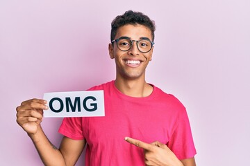 Young handsome african american man holding omg message paper smiling happy pointing with hand and...