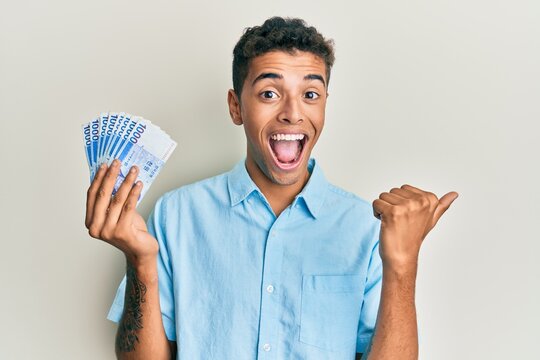 Young handsome african american man holding 1000 south korean won banknotes pointing thumb up to the side smiling happy with open mouth