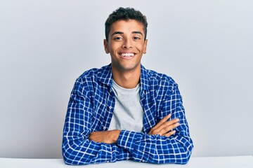 Young handsome african american man wearing casual clothes sitting on the table happy face smiling with crossed arms looking at the camera. positive person.