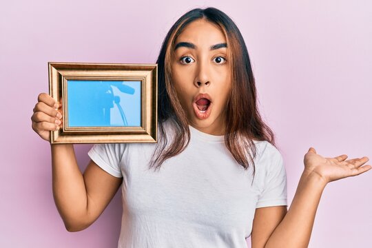 Young latin woman holding empty frame scared and amazed with open mouth for surprise, disbelief face