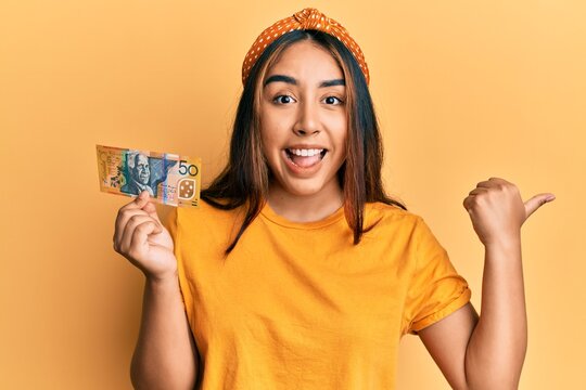 Young latin woman holding 50 australian dollar banknote pointing thumb up to the side smiling happy with open mouth
