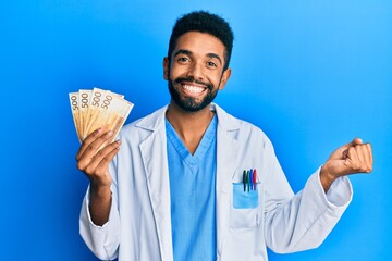 Handsome hispanic man with beard wearing medical uniform holding 500 norwegian krone screaming proud, celebrating victory and success very excited with raised arm