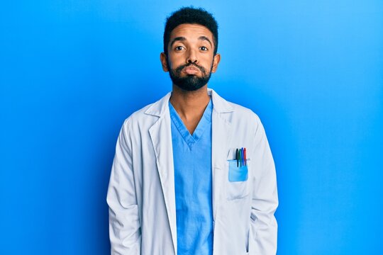 Handsome hispanic man with beard wearing doctor uniform puffing cheeks with funny face. mouth inflated with air, crazy expression.