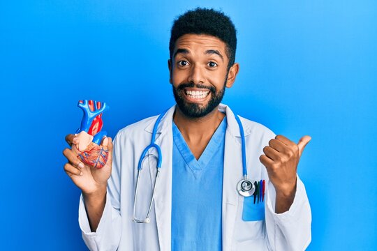 Handsome hispanic man with beard wearing doctor uniform holding heart pointing thumb up to the side smiling happy with open mouth