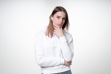 A sweet girl in her 20s with fair hair is pensive and thoughtful listening to music in her white wireless earphones, studio photo