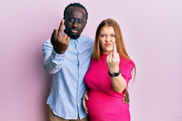Young interracial couple expecting a baby, touching pregnant belly showing middle finger, impolite...