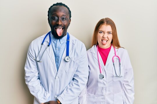 Young interracial couple wearing doctor uniform and stethoscope sticking tongue out happy with funny expression. emotion concept.