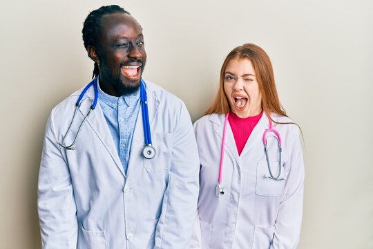 Young interracial couple wearing doctor uniform and stethoscope winking looking at the camera with sexy expression, cheerful and happy face.