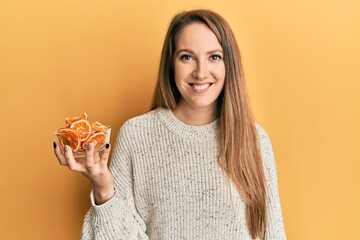 Young blonde woman holding bowl of dry orange looking positive and happy standing and smiling with a confident smile showing teeth