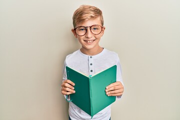 Little caucasian boy kid reading a book wearing glasses smiling with a happy and cool smile on face. showing teeth.