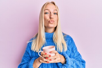 Young blonde girl wearing winter sweater and drinking a cup of hot coffee looking at the camera blowing a kiss being lovely and sexy. love expression.