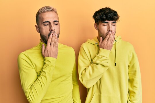 Homosexual gay couple standing together wearing yellow clothes bored yawning tired covering mouth with hand. restless and sleepiness.