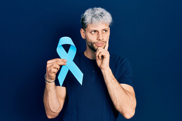 Young hispanic man with modern dyed hair holding pink cancer ribbon serious face thinking about question with hand on chin, thoughtful about confusing idea