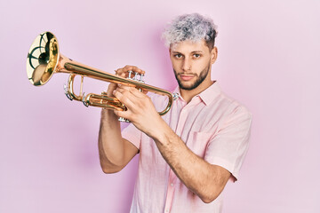 Young hispanic man with modern dyed hair playing trumpet relaxed with serious expression on face....