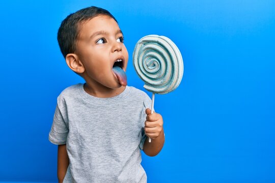 Adorable latin toddler eating delicious lollipop over isolated blue background.