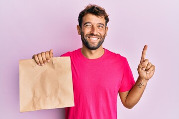 Handsome man with beard holding take away paper bag smiling with an idea or question pointing finger with happy face, number one