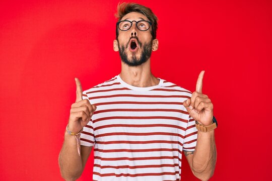 Handsome caucasian man with beard wearing casual clothes and glasses amazed and surprised looking up and pointing with fingers and raised arms.