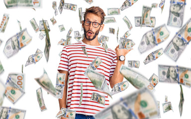 Handsome blond man with beard wearing casual clothes and glasses surprised pointing with hand...
