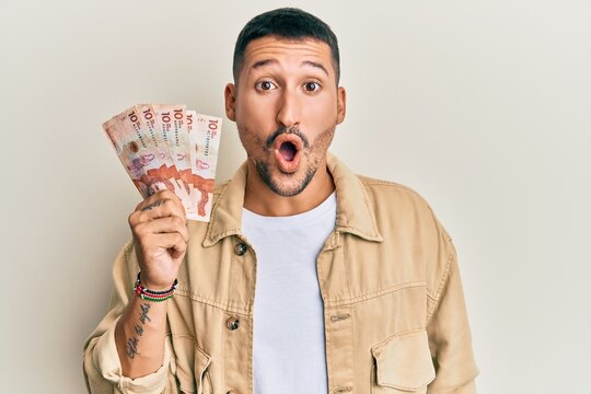 Handsome man with tattoos holding 10 colombian pesos banknotes scared and amazed with open mouth for surprise, disbelief face