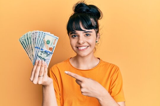 Young hispanic girl holding dollars smiling happy pointing with hand and finger