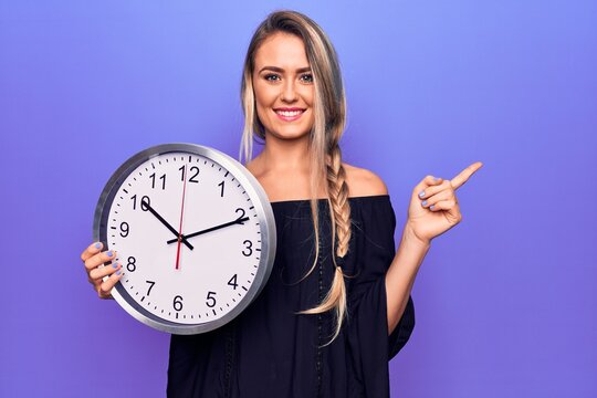 Young beautiful blonde woman doing countdown using big clock over purple background smiling happy pointing with hand and finger to the side