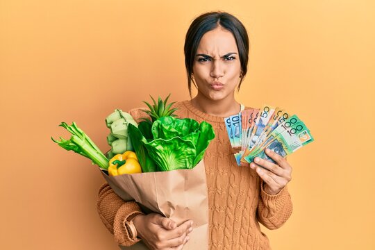 Young brunette woman holding groceries bag and australian dollars banknotes making fish face with mouth and squinting eyes, crazy and comical.