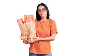 Young beautiful latin woman wearing glasses holding paper bag with bread thinking attitude and sober expression looking self confident